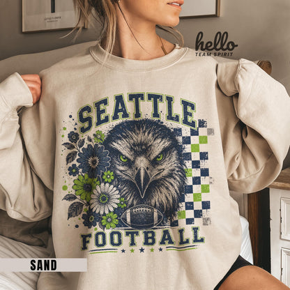 May include: Sand-colored sweatshirt featuring a graphic of a fierce eagle with green eyes, surrounded by blue and green flowers. The words "SEATTLE FOOTBALL" are printed above and below the eagle, with a football graphic. The word "SAND" is printed at the bottom.